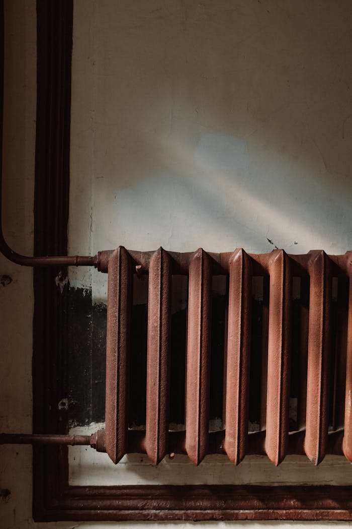 Close-up of a rusty steam radiator with pipes in an old building, casting shadows on the wall.